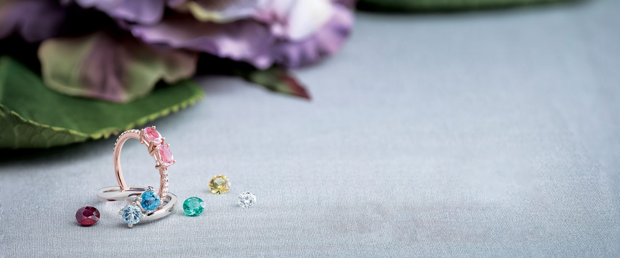 Rose gold ring with pink stones beside loose rubies, sapphires and an emerald on silk